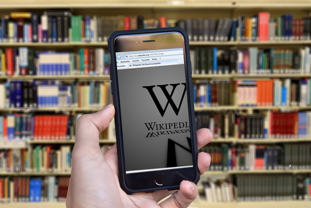 Une bibliothèque en fond d'image, une main avec un smartphone montrant Wikipédia